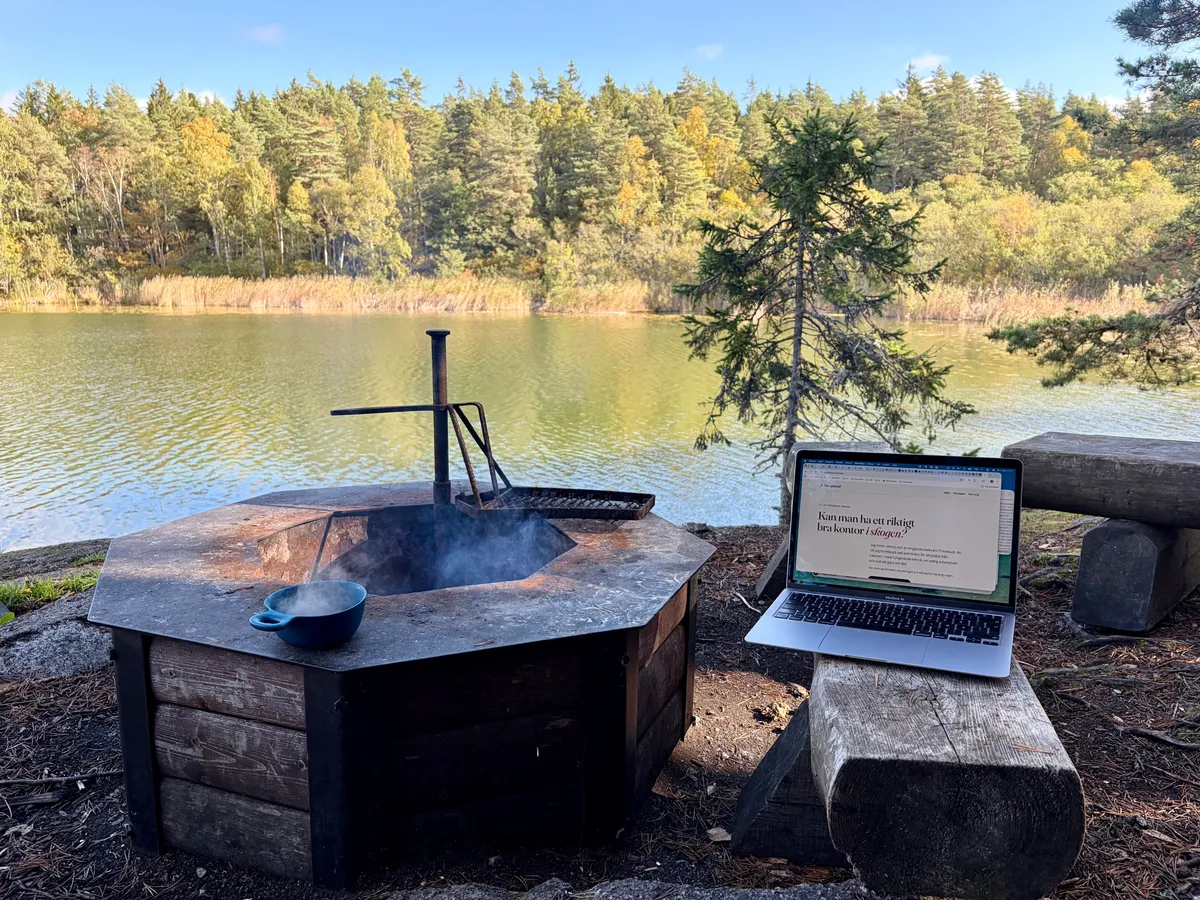 Laptop med skogskod.se på skärmen, på en träbänk bredvid en rykande eldstad vid en sjö omgiven av höstskog.